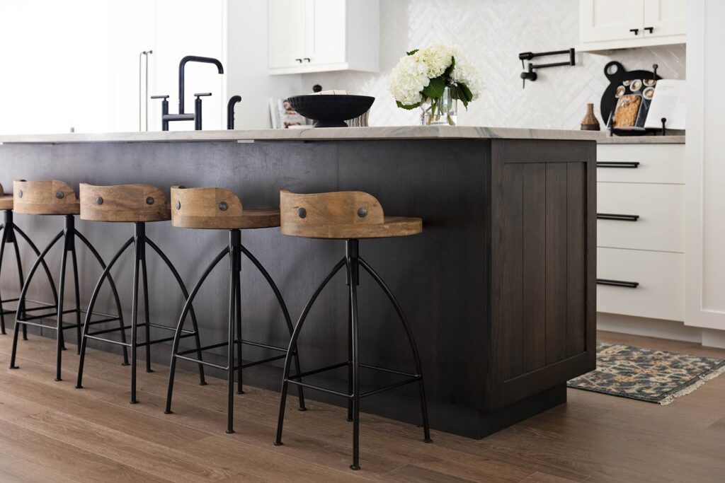 A dark timber kitchen island with a stone worktop and four wooden stools, against white cabinetry and a herringbone tile splashback.