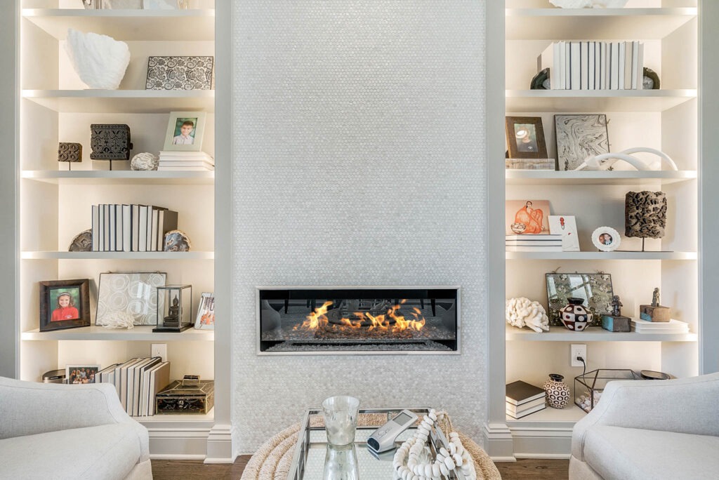 A linear gas fireplace set in a pale tiled chimney breast, flanked by built-in shelving styled with books, art, and objects.