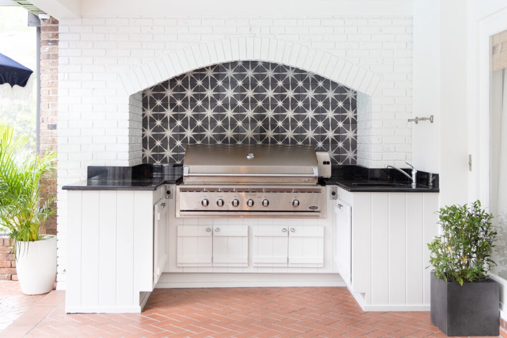 A covered outdoor kitchen in whitewashed brick, with a built-in grill set into a patterned tile alcove, granite counters, and potted plants on a terracotta floor.