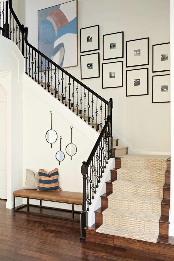An entrance hall staircase with a black wrought-iron balustrade and patterned stair runner, beside a leather bench and a framed gallery wall.